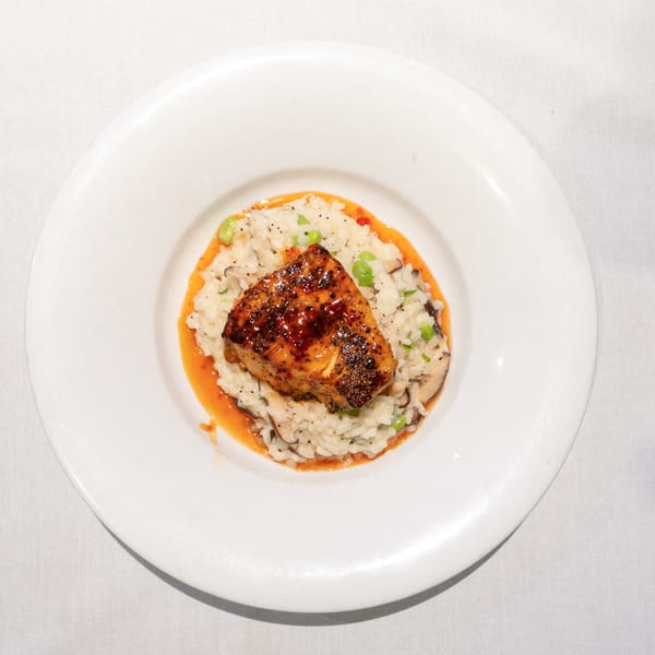 Pan-seared fish over risotto with peas and mushrooms, drizzled with red sauce on a white plate.